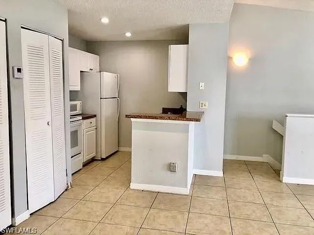 Kitchen featuring white cabinets, dark countertops, a textured ceiling, white appliances, and light tile patterned floors