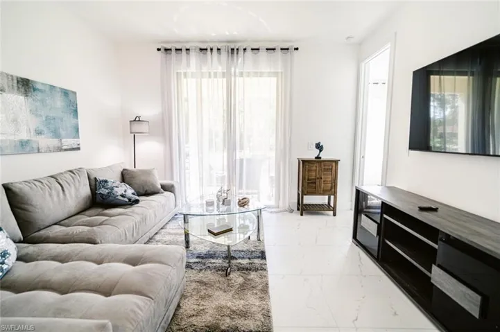 Bright living area featuring large format white tile flooring, a glass sliding door with sheer curtains, recessed lighting, and a wall-mounted television