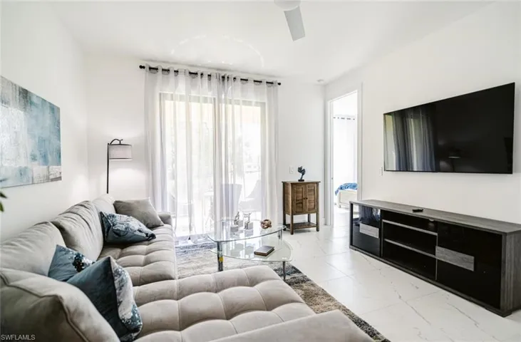 Bright living area featuring white walls, large sliding glass doors with sheer curtains, and light-toned tile flooring