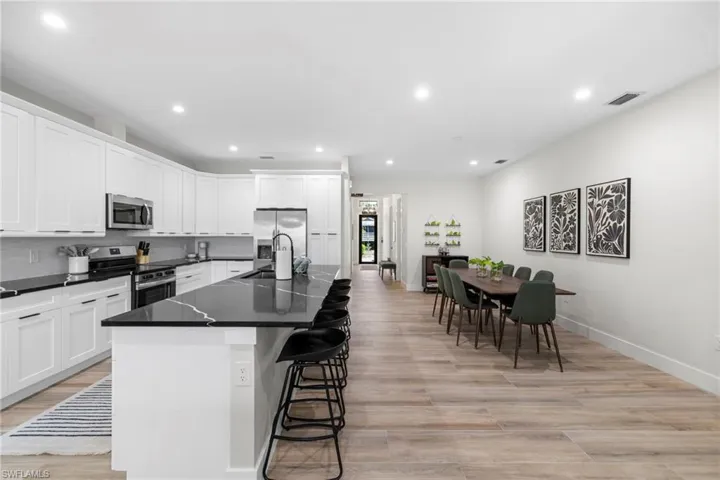 Spacious kitchen and dining area featuring an expansive island with seating, white shaker cabinetry, stainless steel appliances, dark countertops with light veining, and light wood-finish flooring