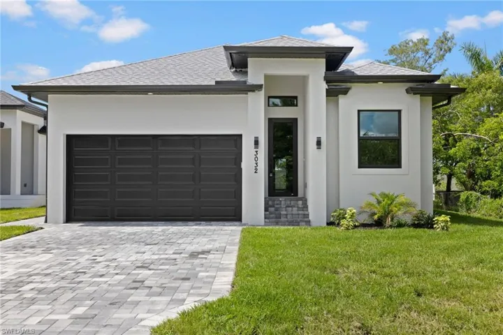 Contemporary stucco exterior featuring a dark paneled garage door, paver driveway, dark-framed windows, and decorative landscaping
