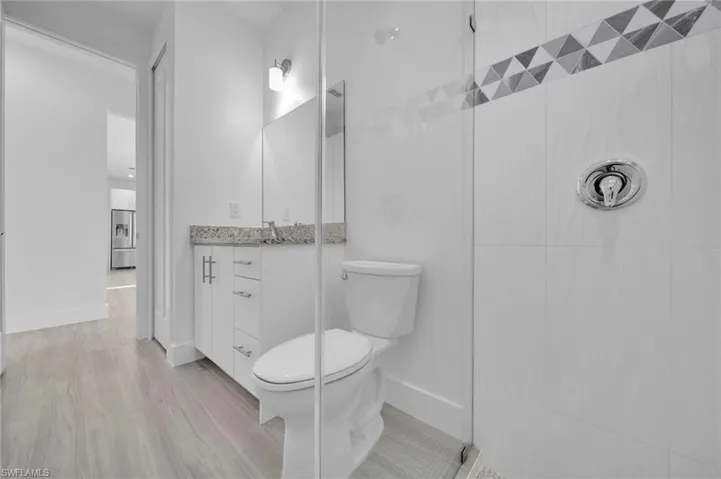 Contemporary bathroom featuring a glass-enclosed shower, white tile surround with decorative accent, a white vanity with granite-finish countertop, and wood-finish flooring