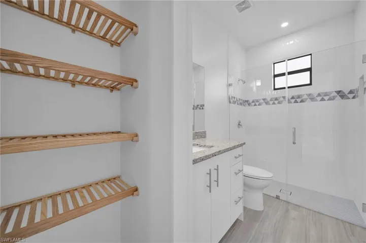 White vanity with a stone-finish countertop, wood-finish shelving, an enclosed shower with a mosaic tile accent, and light wood-finish flooring