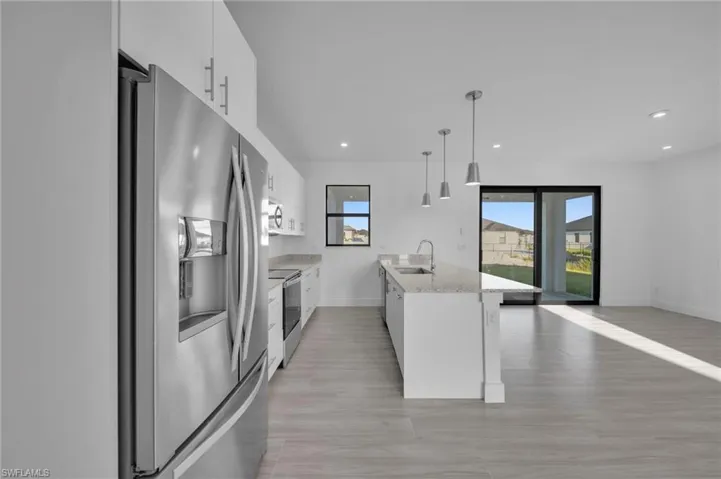 Modern kitchen featuring a stainless steel refrigerator, white cabinetry, a kitchen island with a sink, pendant lighting, and light-toned flooring
