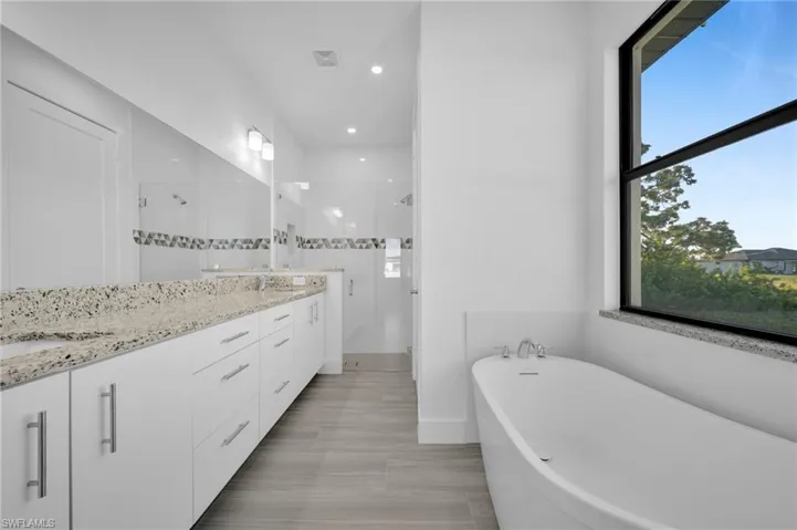 Double vanity with light-toned countertop, wood-finish flooring, freestanding soaking tub, and walk-in shower with glass enclosure