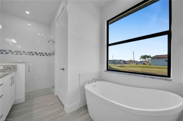 Freestanding soaking tub, glass-enclosed shower with geometric tile inlay, wood-finish flooring, and white cabinetry with granite countertops