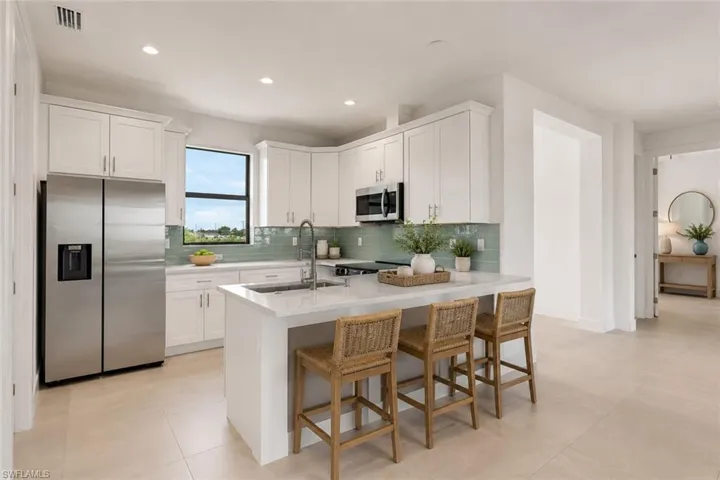 Modern kitchen featuring white cabinetry, a light-toned countertop island with seating, a stainless steel refrigerator, and a light blue tile backsplash