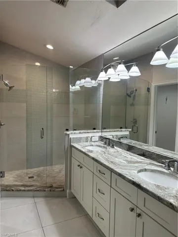 Spa-inspired primary bathroom featuring a frameless glass-enclosed shower with marble-look tile surround, dual-sink vanity with polished stone countertops, chrome fixtures, and recessed and pendant lighting throughout