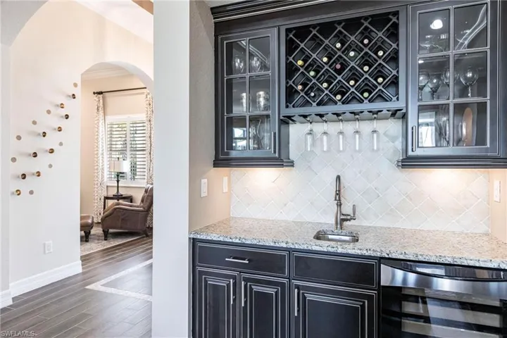 Indoor wet bar featuring light stone countertops, dark cabinetry, beverage cooler, arched walkways, and ornamental molding
