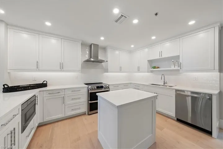 Kitchen with tasteful backsplash, light wood-type flooring, wall chimney exhaust hood, sink, and appliances with stainless steel finishes