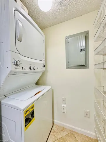 Washroom featuring electric panel, a textured ceiling, stacked washer / drying machine, and light tile patterned floors