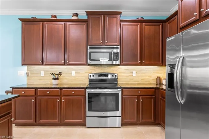 Kitchen featuring stainless steel appliances, decorative backsplash, dark stone countertops, crown molding, and light tile patterned floors