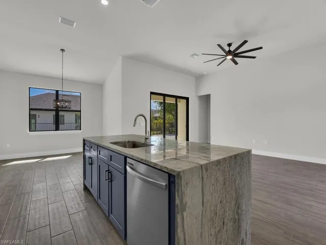 Kitchen with dark stone countertops, wood finish floors, stainless steel dishwasher, a center island with sink, and pendant lighting