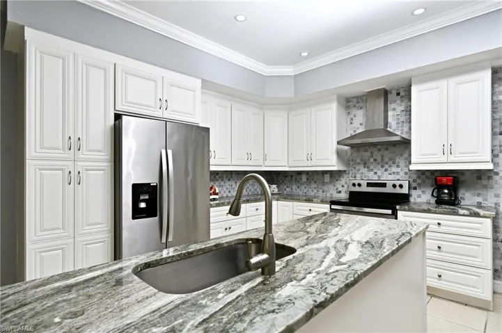Kitchen featuring wall chimney range hood, stainless steel appliances, light tile patterned floors, stone counters, and white cabinets