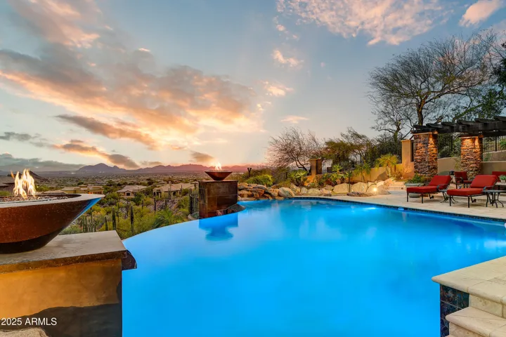 As night falls, the glowing fire bowls cast reflections across the infinity pool, accentuating the drama of the mountain views. This serene space offers an exquisite backdrop for twilight entertaining in this Scottsdale luxury home.