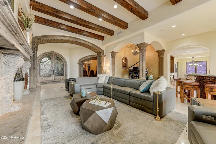 A sophisticated living space with wood ceiling beams, ornate stone fireplace, and inviting sectional seating. This open-concept area blends classic architecture with modern comfort, perfect for hosting or unwinding in style.