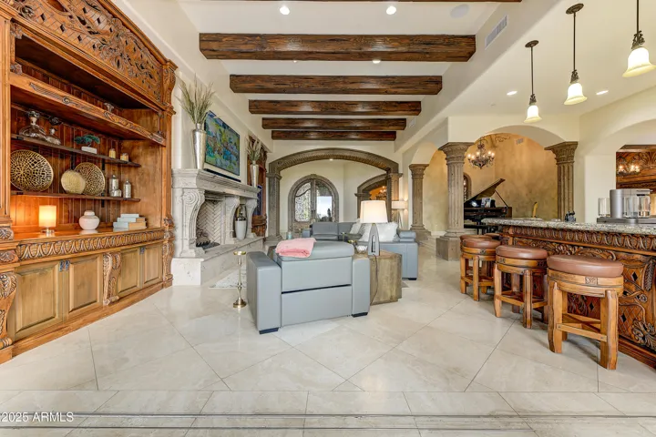 The grand living area offers seamless indoor entertaining with a spacious open floor plan, beamed ceilings, and a beautifully carved bar. Natural stone flooring and artistic archways define this upscale desert retreat.