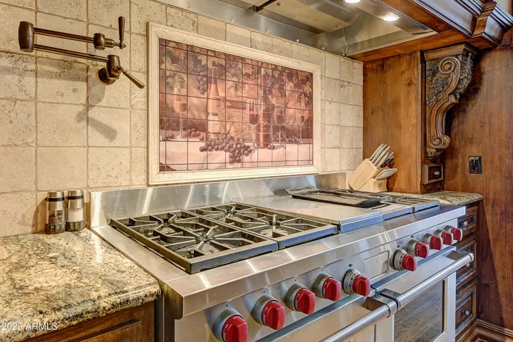 This chef’s kitchen boasts a Wolf gas range with red knobs, custom wood hood, granite counters, pot filler, and a tile backsplash with vineyard mural—a luxurious setting for culinary creativity.