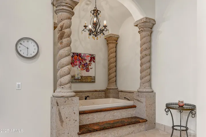 A sunken jetted tub sits regally between custom stone columns and beneath a romantic chandelier. This spa-inspired alcove is the ultimate place to unwind and embrace luxury living with high-end finishes and timeless design.