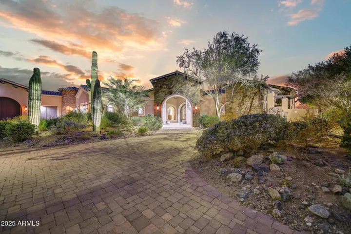 A captivating front elevation with desert landscaping, towering saguaro cacti, stone accents, and a grand arched entryway that welcomes you into this exquisite Scottsdale estate nestled along the Via Linda corridor.