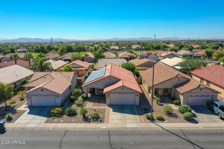 This elevated street-level aerial showcases the home’s attractive curb appeal in Buckeye, featuring a tile roof, desert landscaping, and a clean driveway approach. The surrounding neighborhood reflects well-maintained homes and a quiet residential setting, ideal for buyers seeking a well-established community with energy-efficient design elements visible throughout the area.
