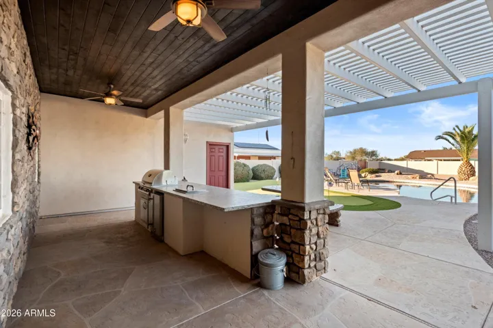Spacious covered patio designed for outdoor living and entertaining, featuring a built-in barbecue, compact refrigerator, integrated ice chest, and generous counter space. Ceiling fans and shade coverage create a comfortable space with direct views of the pool and backyard.