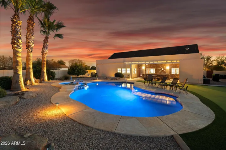 Twilight view of the illuminated pool and spa with landscape lighting, palms, and covered patio, showcasing the backyard’s evening ambiance and resort-style design.