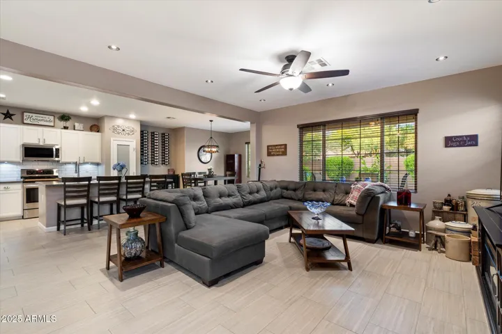 This inviting living room offers wide windows overlooking the backyard, enhancing indoor-outdoor living. Recessed lighting, a ceiling fan, and durable tile flooring add comfort and style, while the open design connects directly to the kitchen for functional flow.