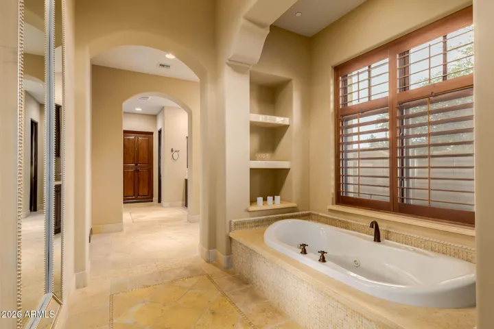 Jetted Tub surrounded with Stone and Travertine, Shelves on both sides and Window with Plantation Shutters.