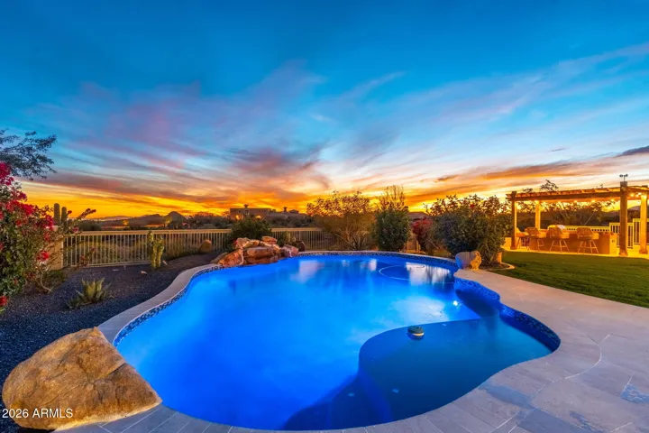 Resort-style Scottsdale backyard showcasing a glowing pool, spa, and fully equipped outdoor kitchen beneath a pergola, ideal for evening entertaining.