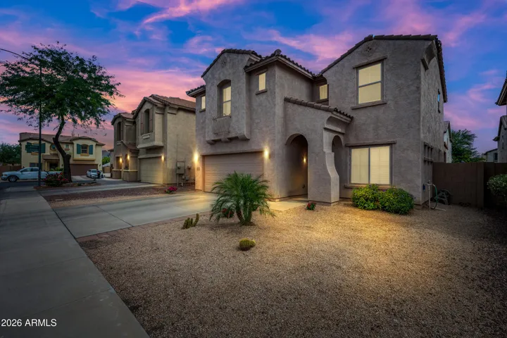 Beautiful twilight exterior showcasing a two-story Arizona home with warm exterior lighting, desert landscaping, and a dramatic pink-and-blue sunset sky. The home features a spacious driveway, attached garage, stucco exterior, tile roof accents, and low-maintenance gravel landscaping, creating inviting curb appeal with a peaceful neighborhood feel.