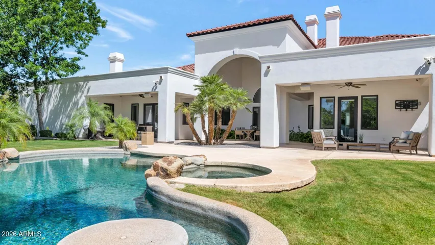 This view captures the full relationship between the home and its outdoor environment — the crisp white Mediterranean facade reflected in the pool's surface, arched covered patios extending the living space on either side, and mature palms framing the scene with effortless Arizona grandeur. Nearly an acre of irrigated grounds surrounds it all.