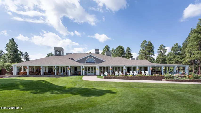 Beautiful clubhouse exterior with manicured green lawns, covered patios, and luxury gathering spaces surrounded by scenic pine forest views.