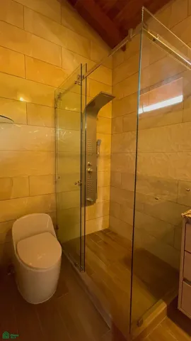 Modern bathroom featuring floor-to-ceiling tile finishes, glass-enclosed rainfall shower, warm wood accents, and elegant contemporary design.
