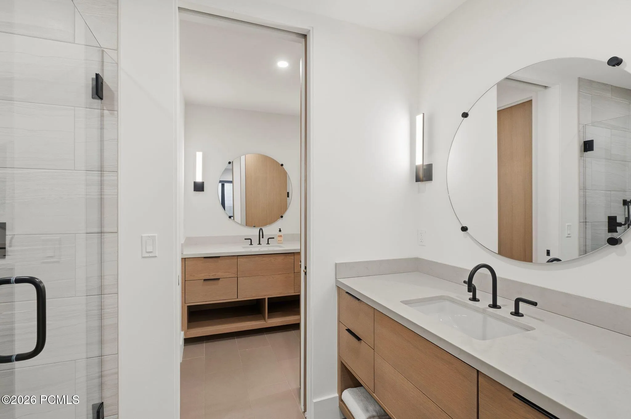 Contemporary bathroom featuring light wood-finish dual vanities with an integrated sink, matte black fixtures, and a large circular mirrors