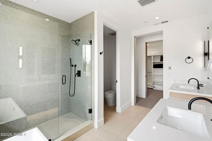 Spacious bathroom featuring a glass-enclosed shower with matte black fixtures, dual vanities with white countertops, and a separate water closet