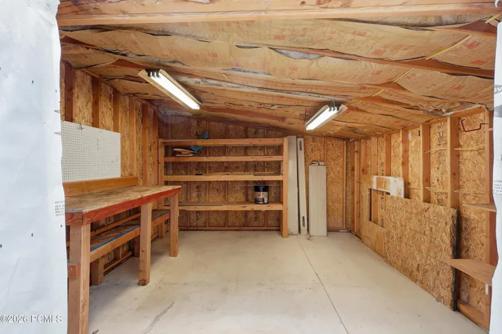 A workshop featuring a 100 amp sub-panel, 220 v outlet, wood-finish wall paneling, built-in wooden shelving, a large workbench with pegboard, and overhead strip lighting.