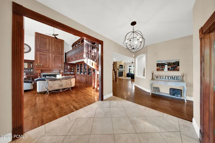 Foyer with tiled flooring and an ornate spherical chandelier, opening to a living area featuring wood-finish flooring, a built-in entertainment center with extensive cabinetry, a fireplace, and an elevated wood-paneled balcony with a carved railing
