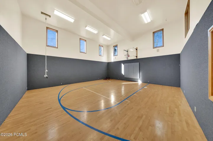 Indoor multi-sport court featuring wood-finish flooring with court markings, padded wall surfaces, a basketball hoop, and multiple windows