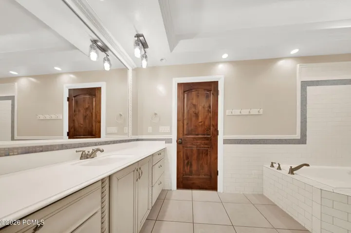 Bathroom featuring a large vanity with dual sinks, an expansive mirror, recessed lighting, a built-in tub with tile surround, and a dark wood-finish interior door