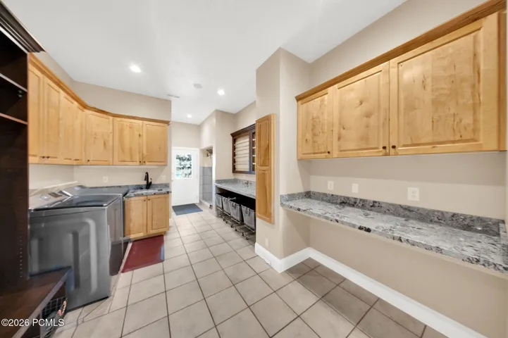 Spacious utility room featuring extensive light wood cabinetry, light-toned tile flooring, and ample counter space with a speckled stone-finish surface