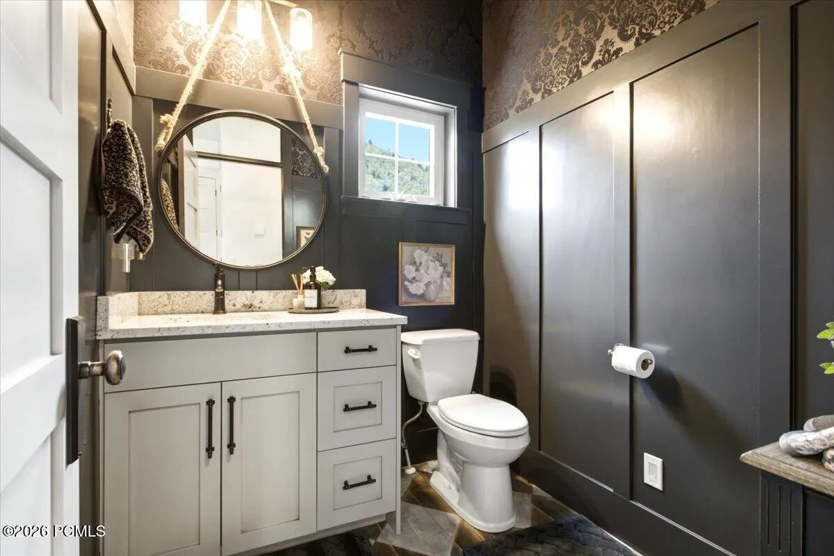 Bathroom featuring a vanity with a light-toned countertop, a round mirror, dark paneled wainscoting, and patterned wallpaper