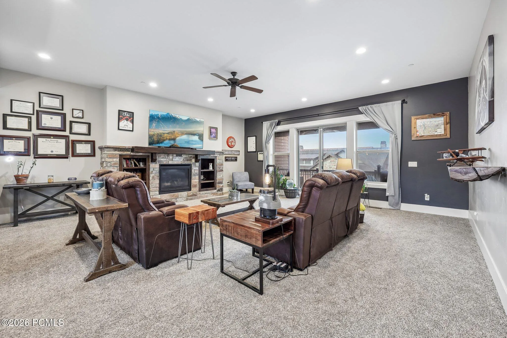 Living room with a stone gas fireplace, ceiling fan and picture windows opening toward the covered deck and Jordanelle Reservoir views.