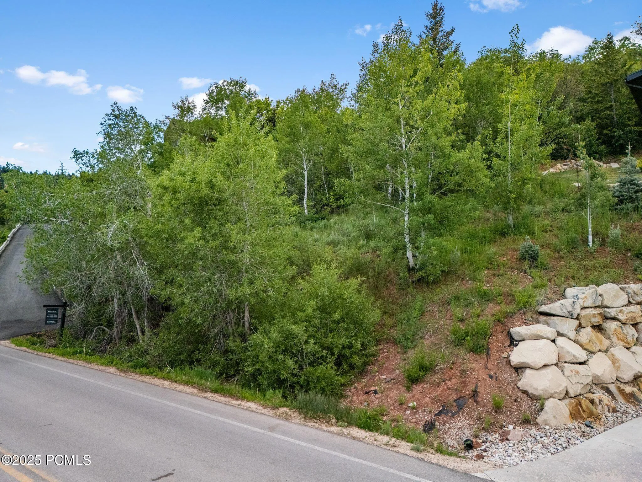 Street-level view of the lot's Stagecoach Drive frontage, with a boulder retaining wall, mature aspens and pines, and the building site rising above the road.