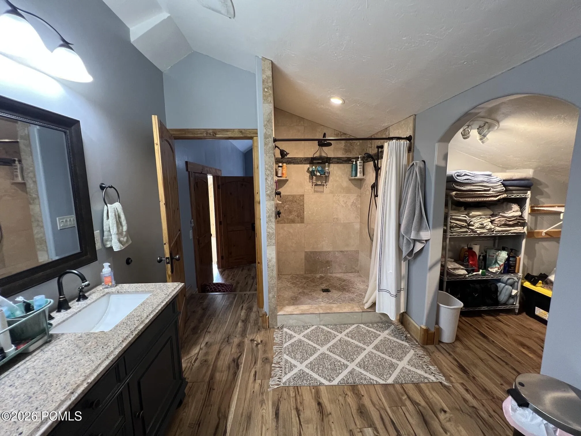Bathroom featuring a double vanity with stone countertop, wood-finish flooring, a tiled shower enclosure, and vaulted ceilings, large walk-in closet to the right.