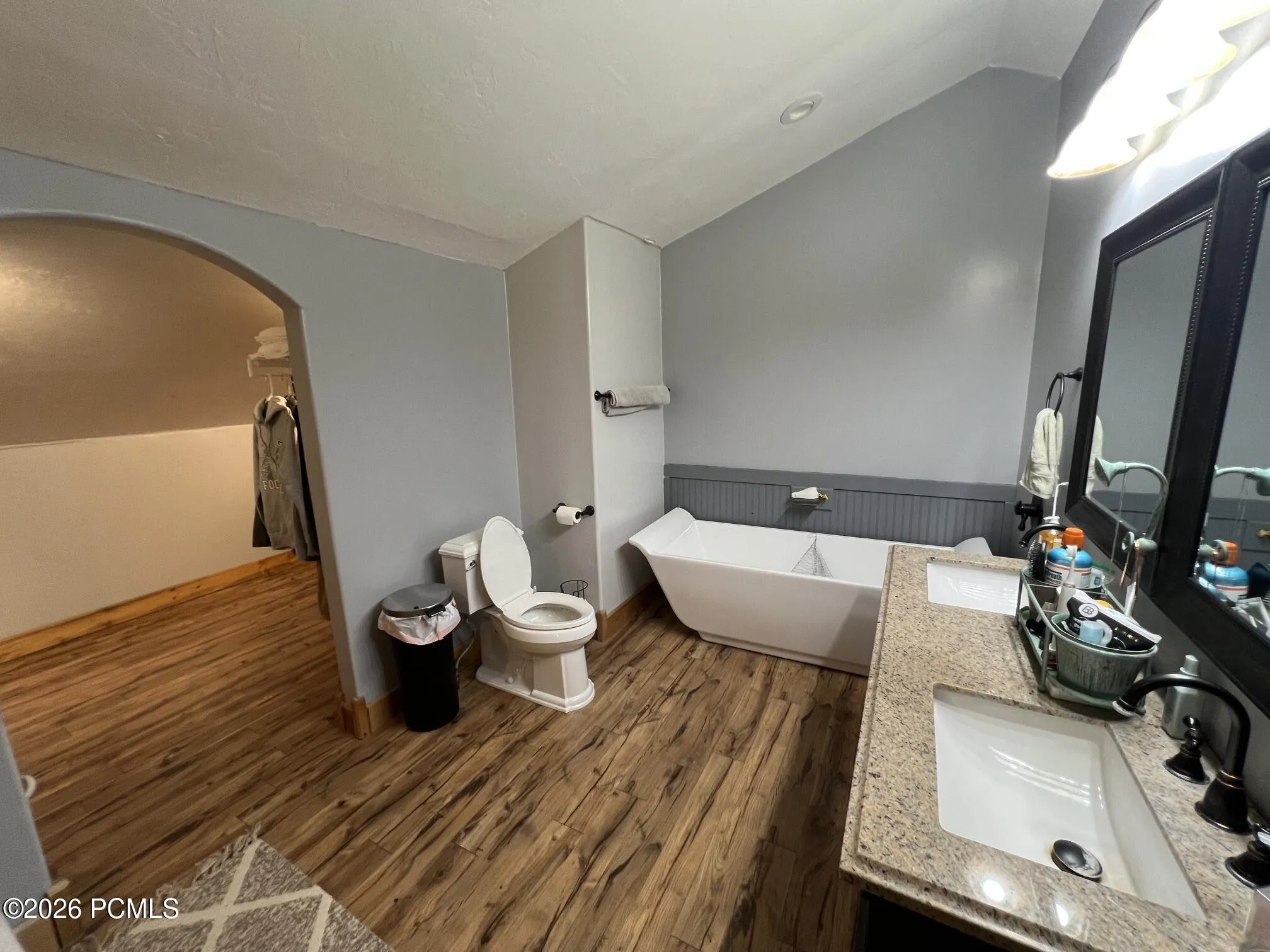 Freestanding soaking tub, dual vanity with granite countertops, wood-finish flooring, and an arched doorway into large walk-in closet