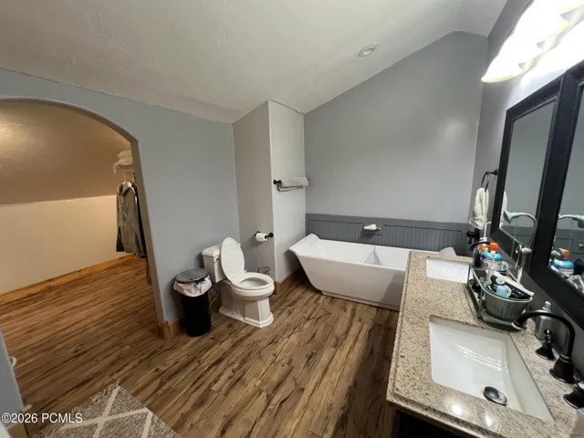 Freestanding soaking tub, dual vanity with granite countertops, wood-finish flooring, and an arched doorway into large walk-in closet