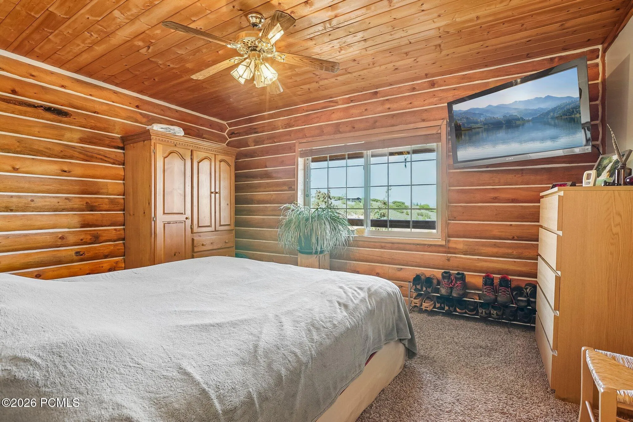 The primary bedroom keeps the log walls and adds a mounted television and a mountain view from bed. Timber Lakes at its most persuasive.The primary bedroom keeps the log walls and adds a mounted television and a mountain view from bed. Timber Lakes at its most persuasive.Primary bedroom with log walls, a ceiling fan, wall-mounted television, and a window with mountain views in Timber Lakes.
