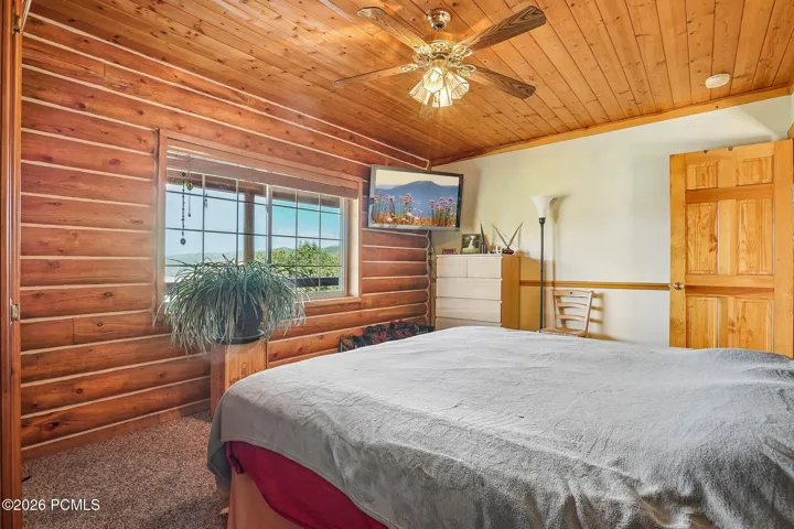A second bedroom with a ceiling fan and windows looking out across the meadow, comfortable enough that guests stay longer than planned.A second bedroom with a ceiling fan and windows looking out across the meadow, comfortable enough that guests stay longer than planned.Bedroom with log walls, a ceiling fan, dresser, and windows framing meadow views.