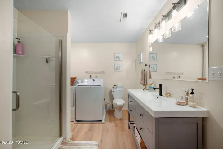 Shower with glass enclosure, single vanity with integrated sink, wood-finish flooring, and wall-mounted lighting